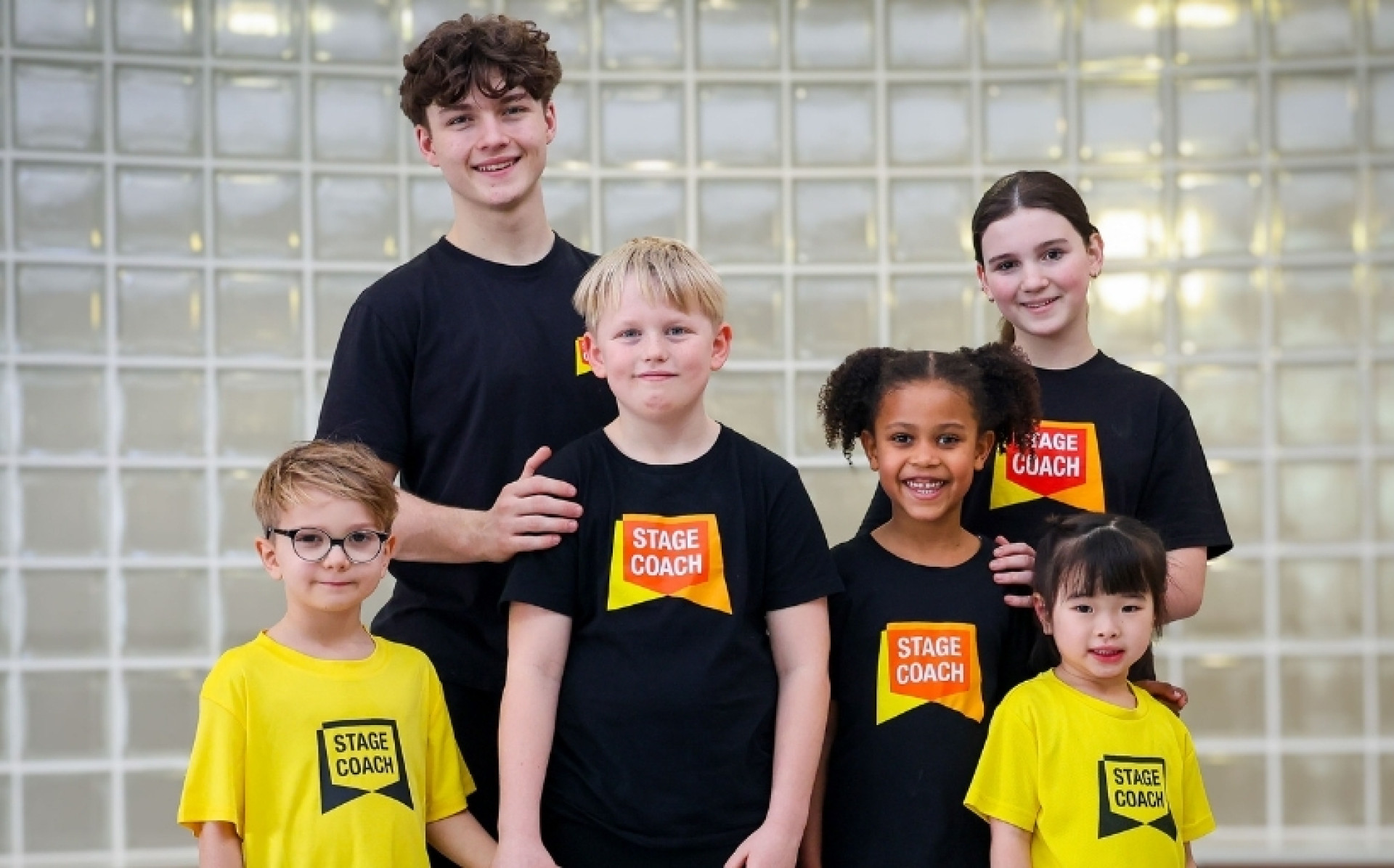 A group of Stagecoach Performing Arts students of different ages smiling together in yellow and black Stagecoach T-shirts